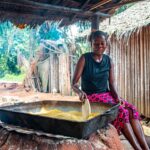 A woman smiles as she prepares cornmeal outdoors in a traditional village setting, capturing cultural authenticity.