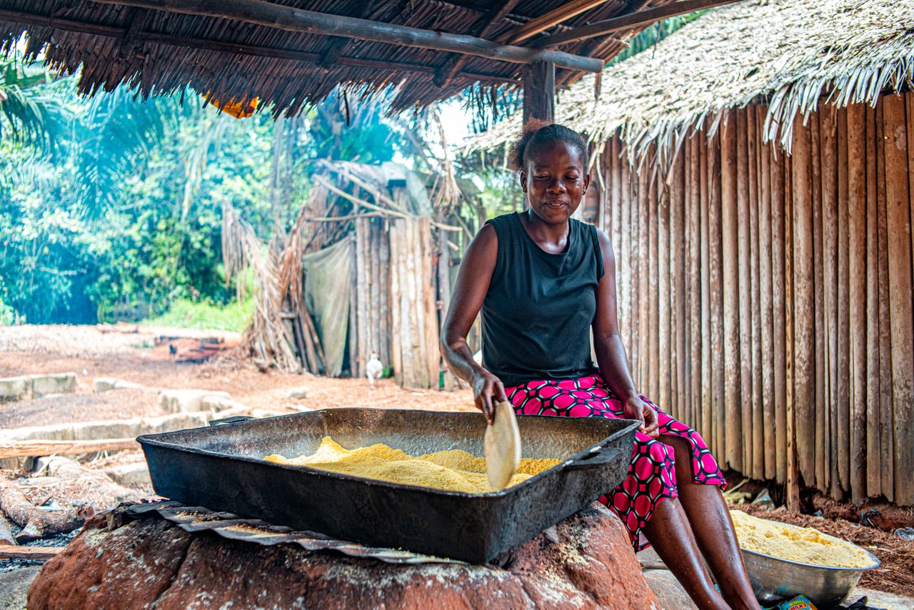 A woman smiles as she prepares cornmeal outdoors in a traditional village setting, capturing cultural authenticity.