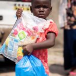A young African girl holding relief supplies in plastic bags outdoors.